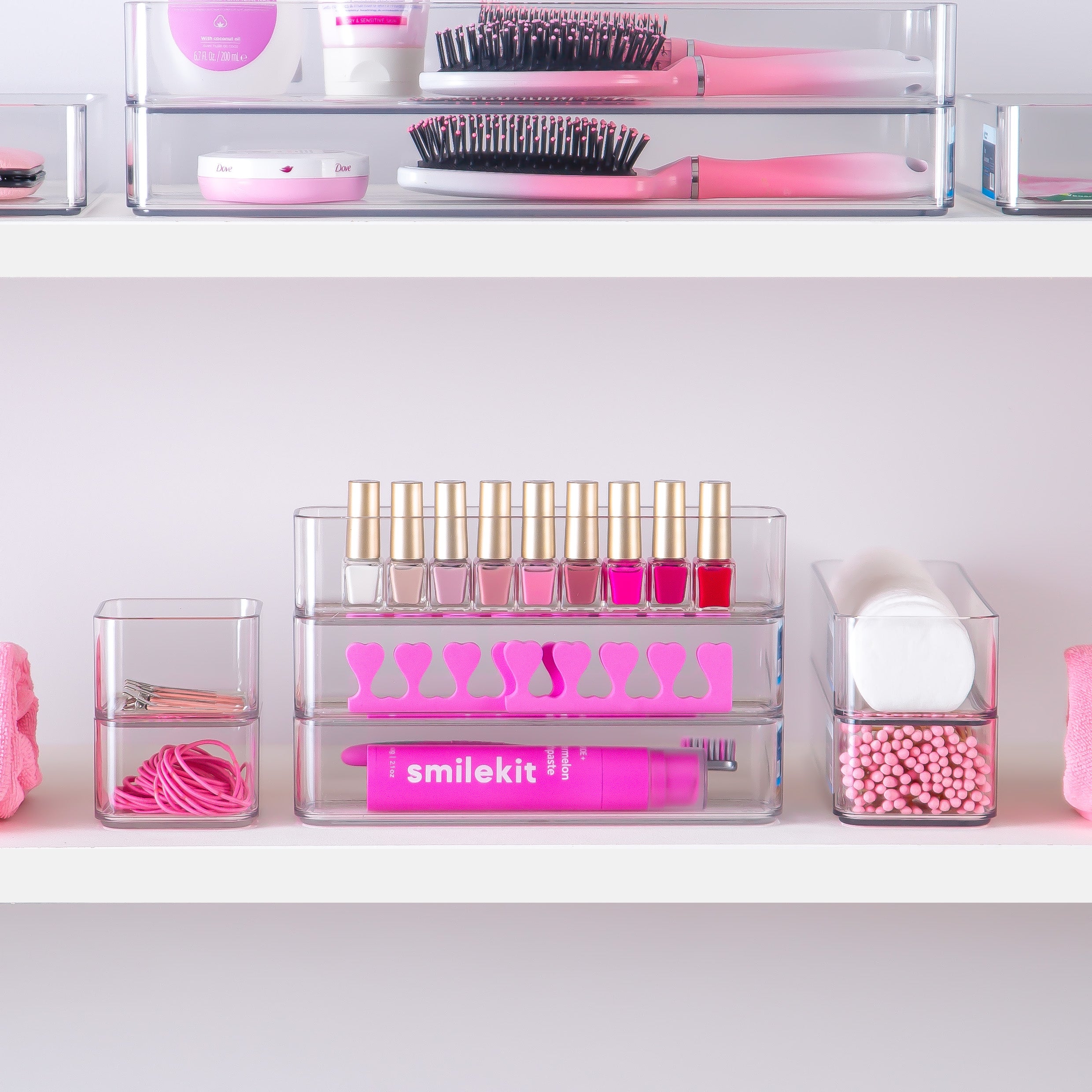 Collection of pink beauty products including hairbrushes, lipsticks, and nail polish on a white shelf in clear storage containers.